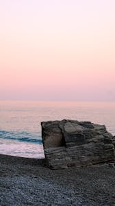 Serene beach landscape with a large rock, calm waves, and pastel sky at sunset.
